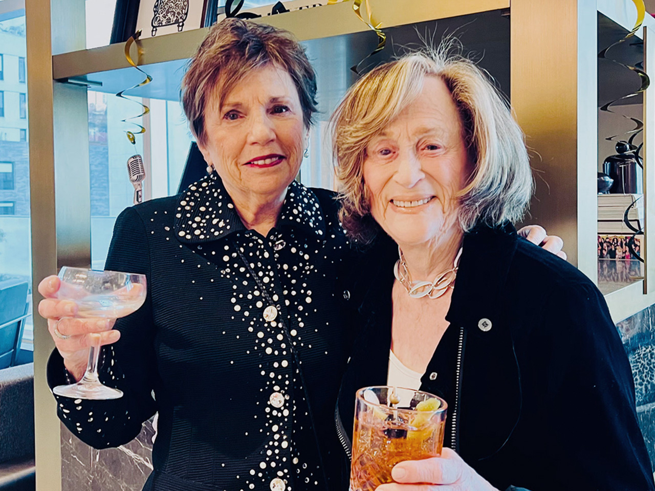 Two smiling women holding drinks, one with a cocktail and the other with champagne, standing indoors with festive decorations behind them.