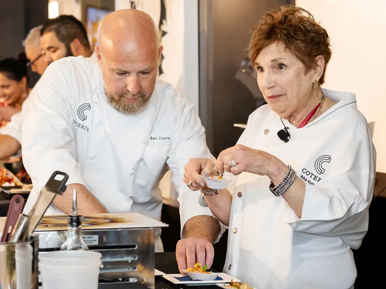 Two chefs in white uniforms prepare dishes together, focusing intently. A man plates food while a woman holds a small dish. Cooking tools visible.