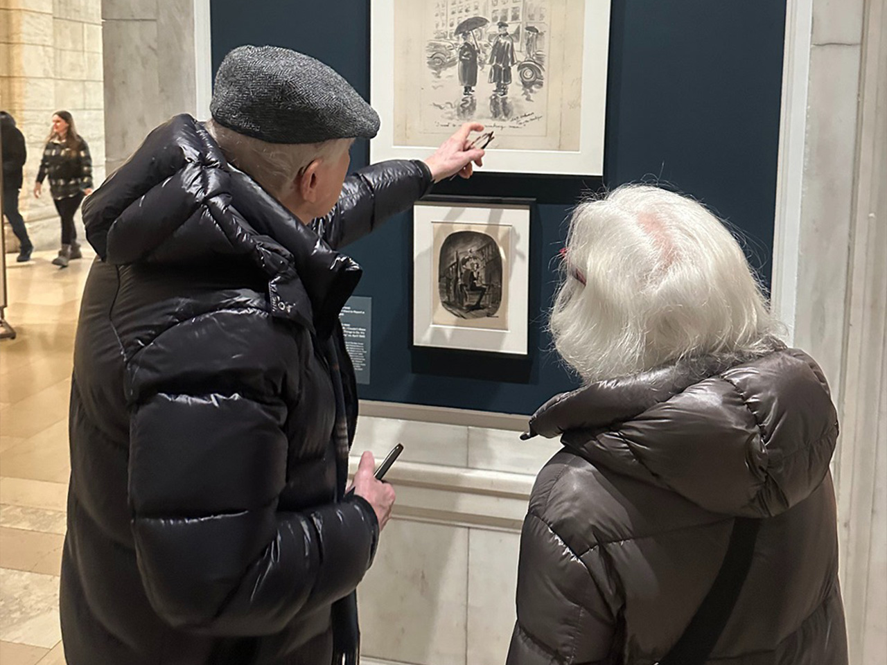 Two people in winter coats and hats observe framed art in a gallery, one pointing at a sketch, the other holding a device.