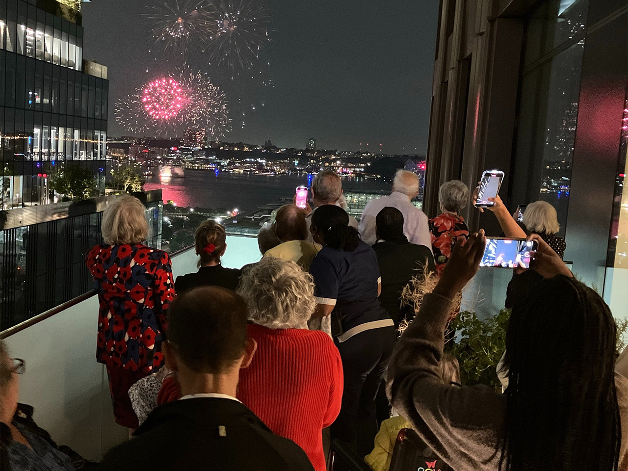 People on a balcony watching a vibrant fireworks display over a cityscape at night, with many capturing the scene on their phones.