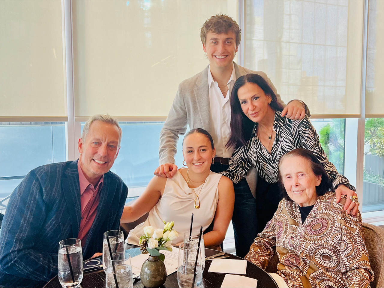 Family smiling at a dining table with glasses and flowers, inside a brightly lit room with a window backdrop.