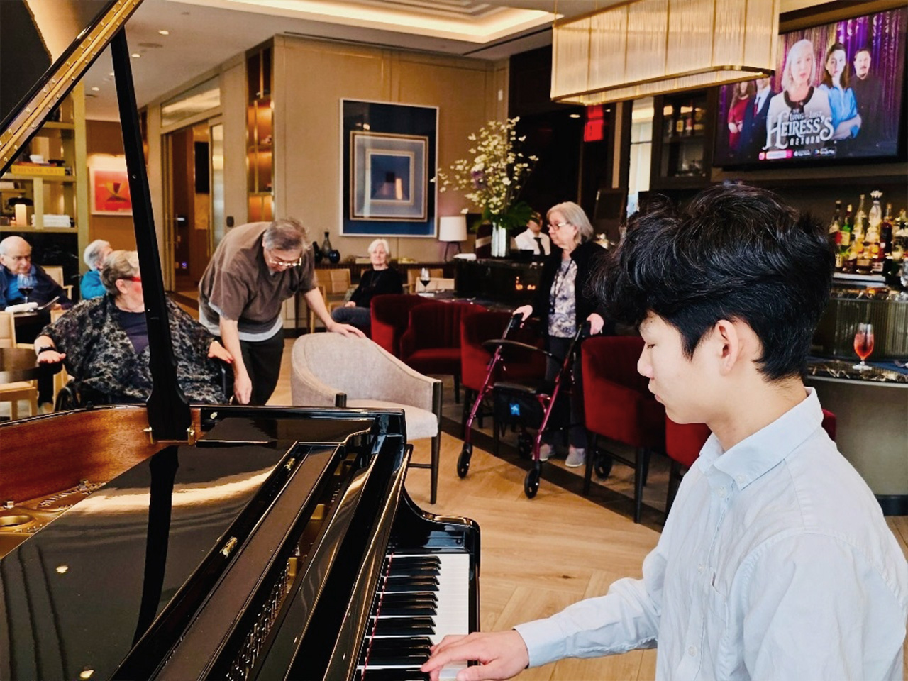 A young person plays the piano in a cozy room with an audience of senior residents seated and standing, a TV visible in the background.