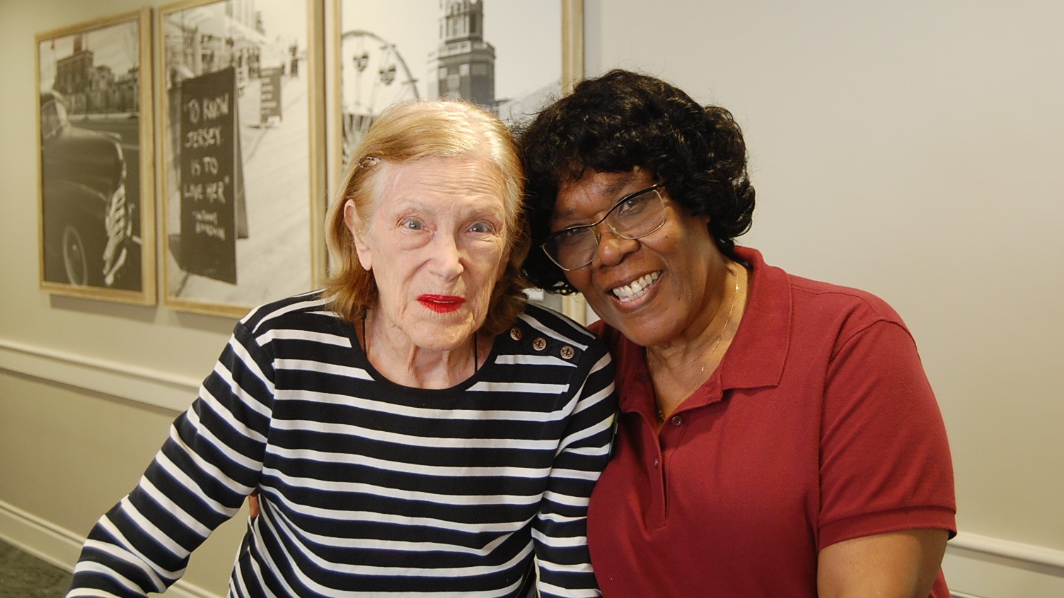 Two women smiling, standing together indoors. One wears a striped shirt, the other a red polo. Black and white framed photos hang in the background.