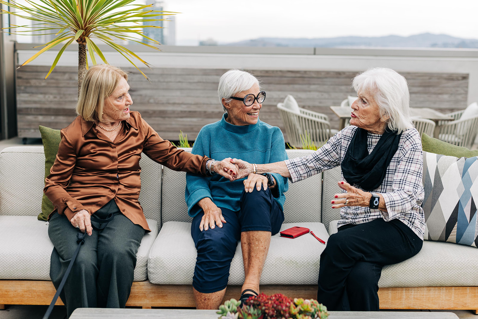 Three elderly women sit on a rooftop patio, engaging in animated conversation and holding hands, with a small plant in the background.