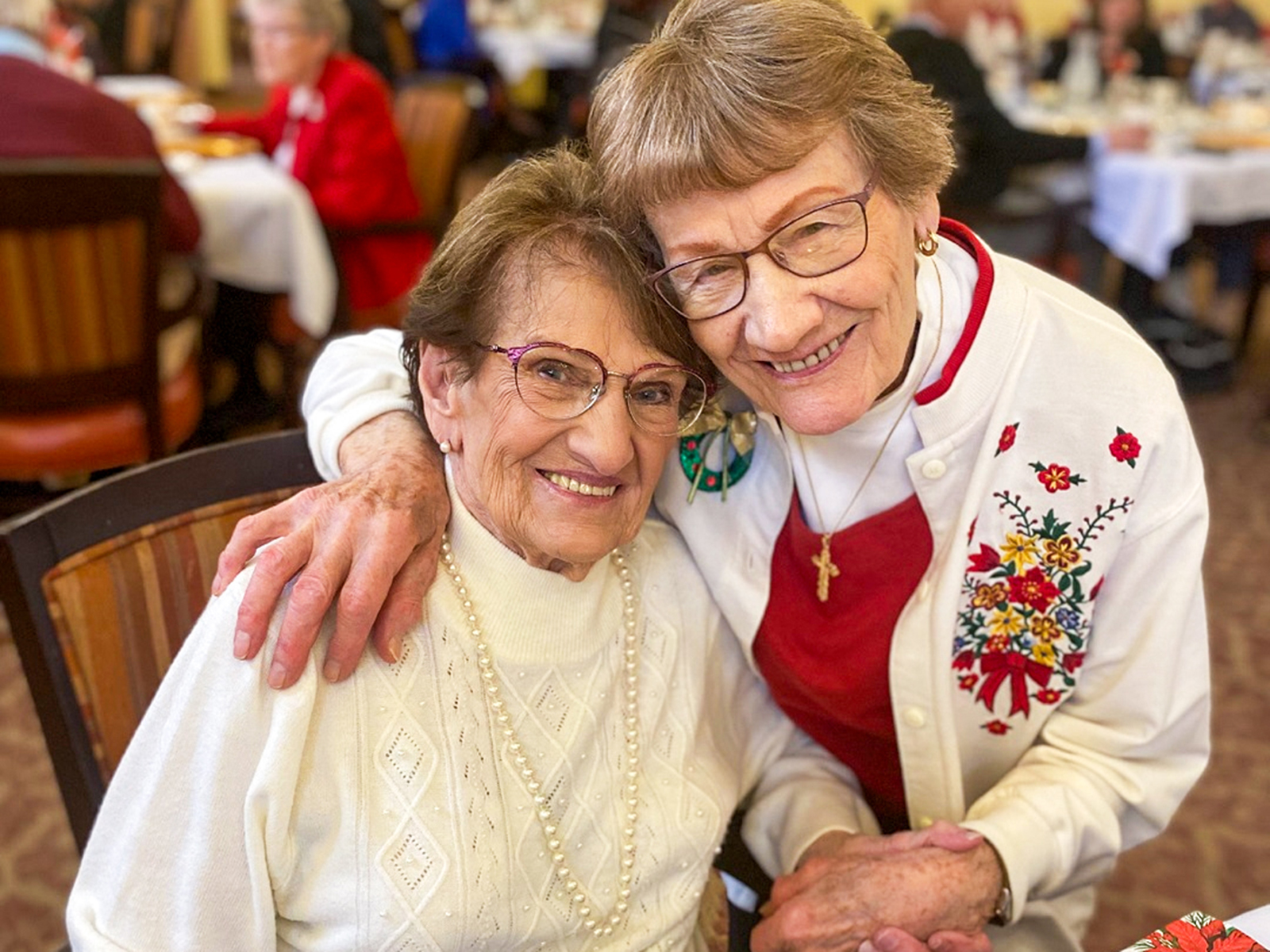 Two older women sit closely together in a dining hall, smiling warmly at the camera. One has her arm around the other, and both wear glasses and festive outfits—one in a white sweater with pearls, the other in a white cardigan with colorful embroidered flowers and a cross necklace. Other people are visible in the softly blurred background.