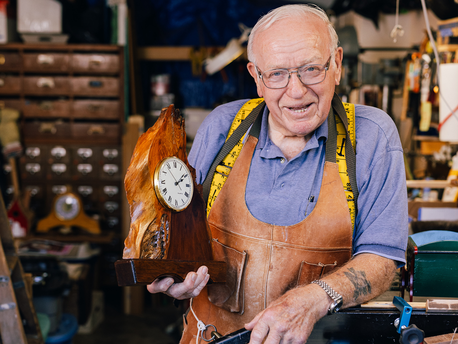 An elderly man in a workshop, wearing glasses, a leather apron, and a tape measure, holding a wooden clock with a Roman numeral face.