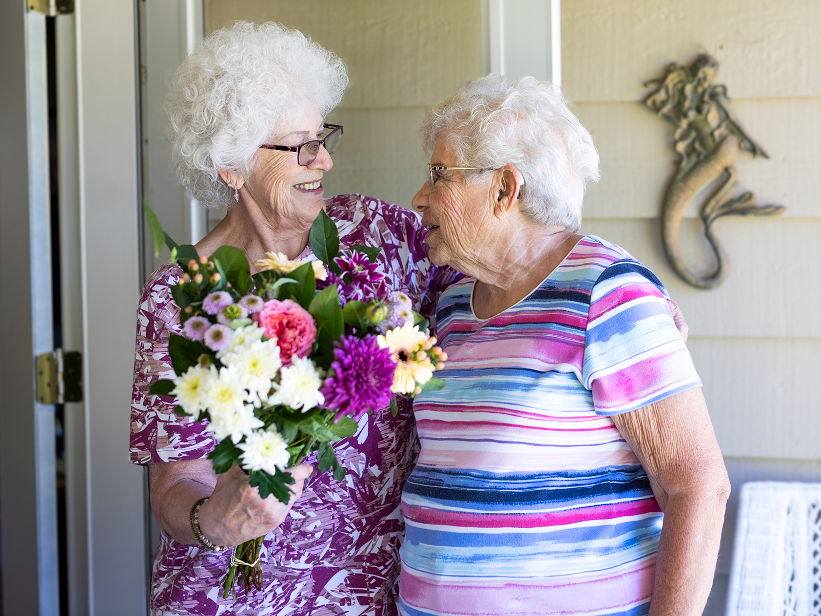 Two elderly women smile at each other, one holding a colorful bouquet of flowers, standing by an open door with a mermaid wall decoration.
