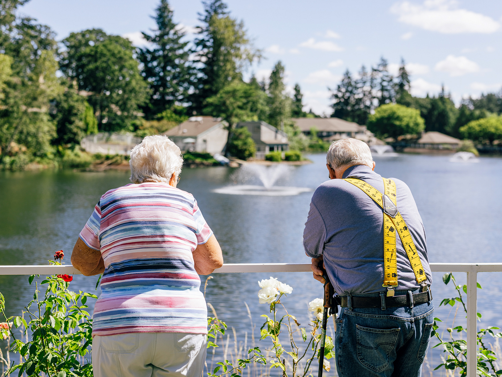 Two elderly people, viewed from behind, lean on a railing overlooking a serene lake with fountains and trees in the background.