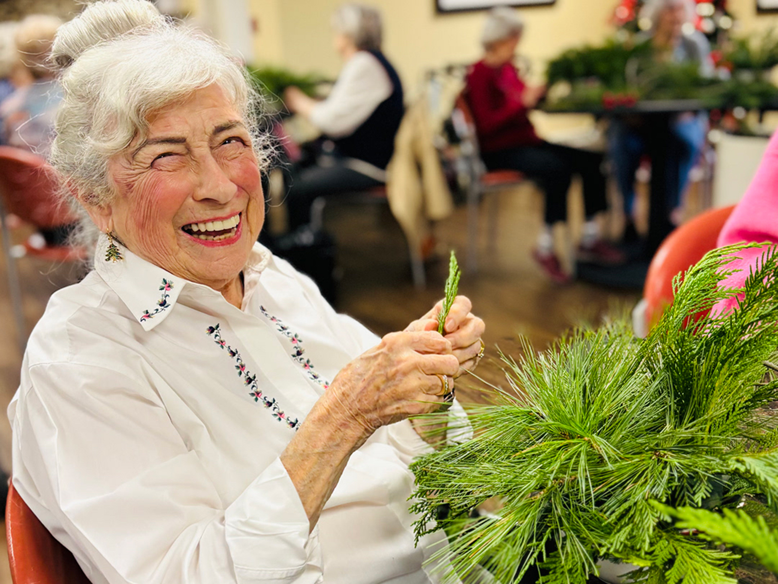 An older woman with white hair in a bun smiles brightly while making a holiday greenery arrangement. She wears a white blouse with floral embroidery and small Christmas-tree earrings. She holds a sprig of evergreen in her hands, with a bundle of greenery in front of her. Other people working on similar crafts are visible in the softly blurred background.