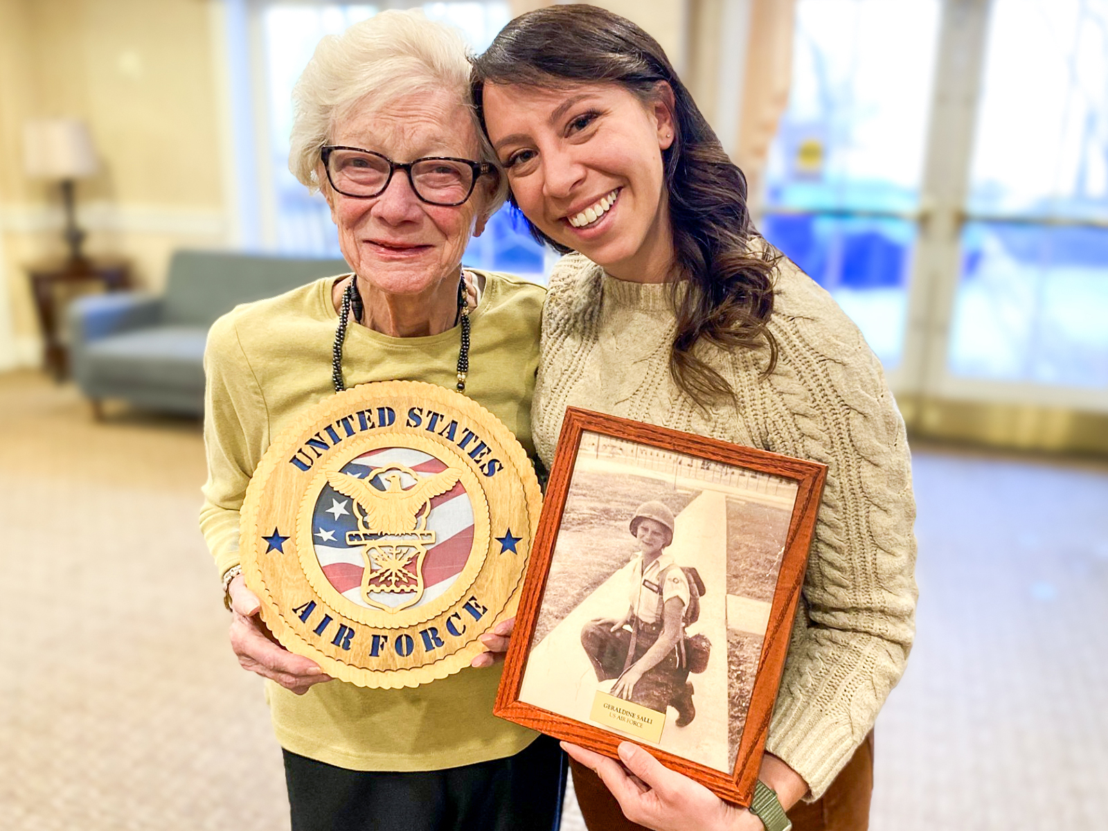 Older woman holding a U.S. Air Force emblem stands with a younger woman holding a framed photo of a man in uniform. Both are smiling.