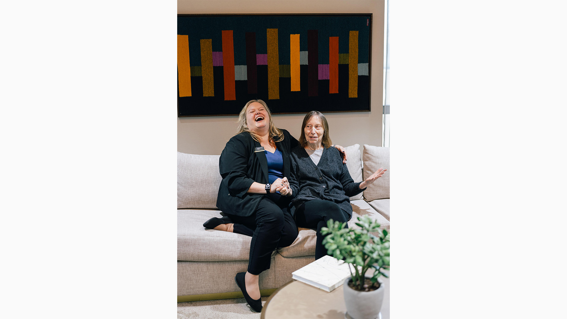 Two women joyfully laughing together on a beige couch. A colorful geometric artwork hangs on the wall behind them. A plant and book are on the table.