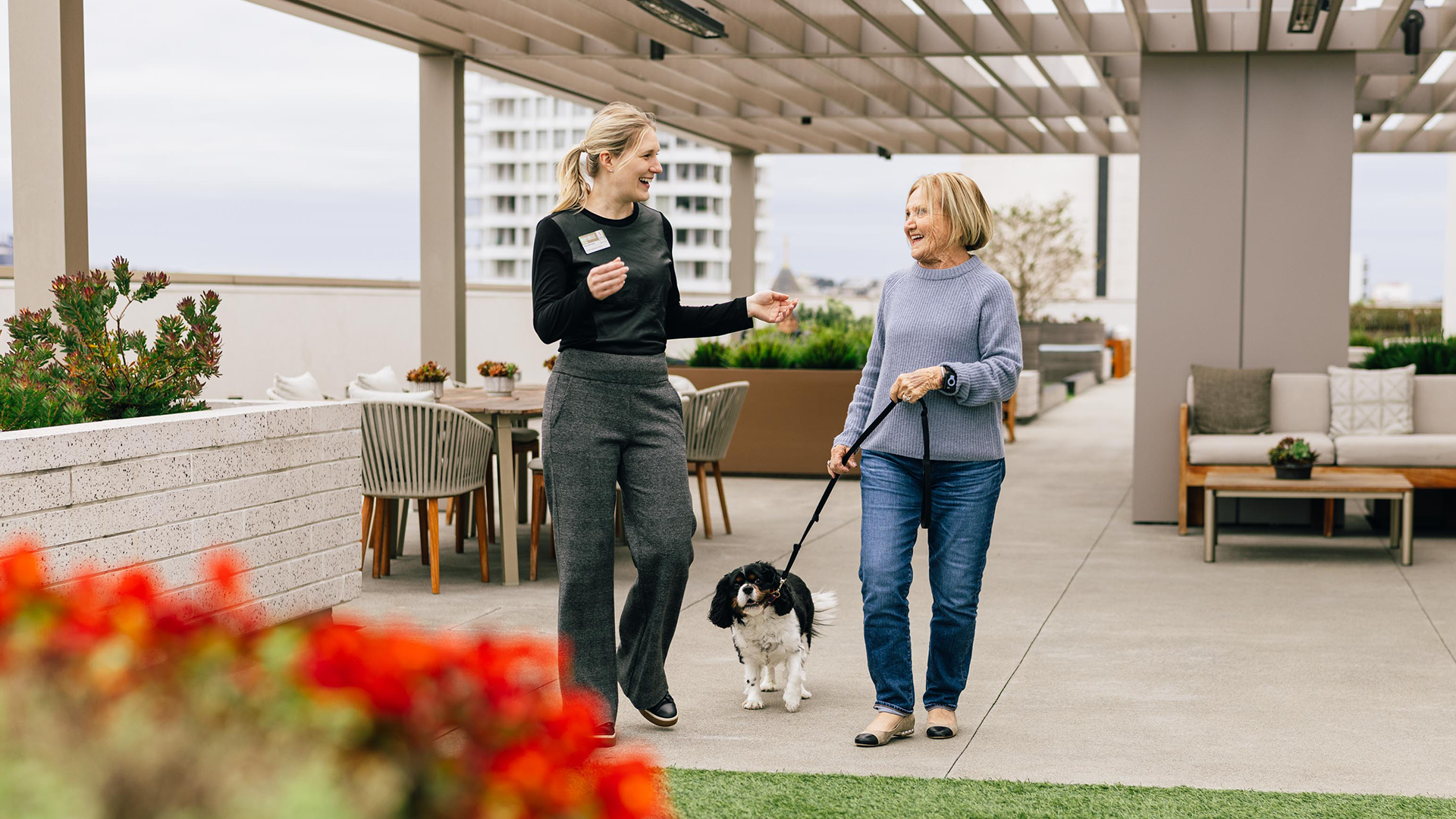 Two women walking and talking on a terrace with a small dog on a leash, surrounded by outdoor furniture and plants.