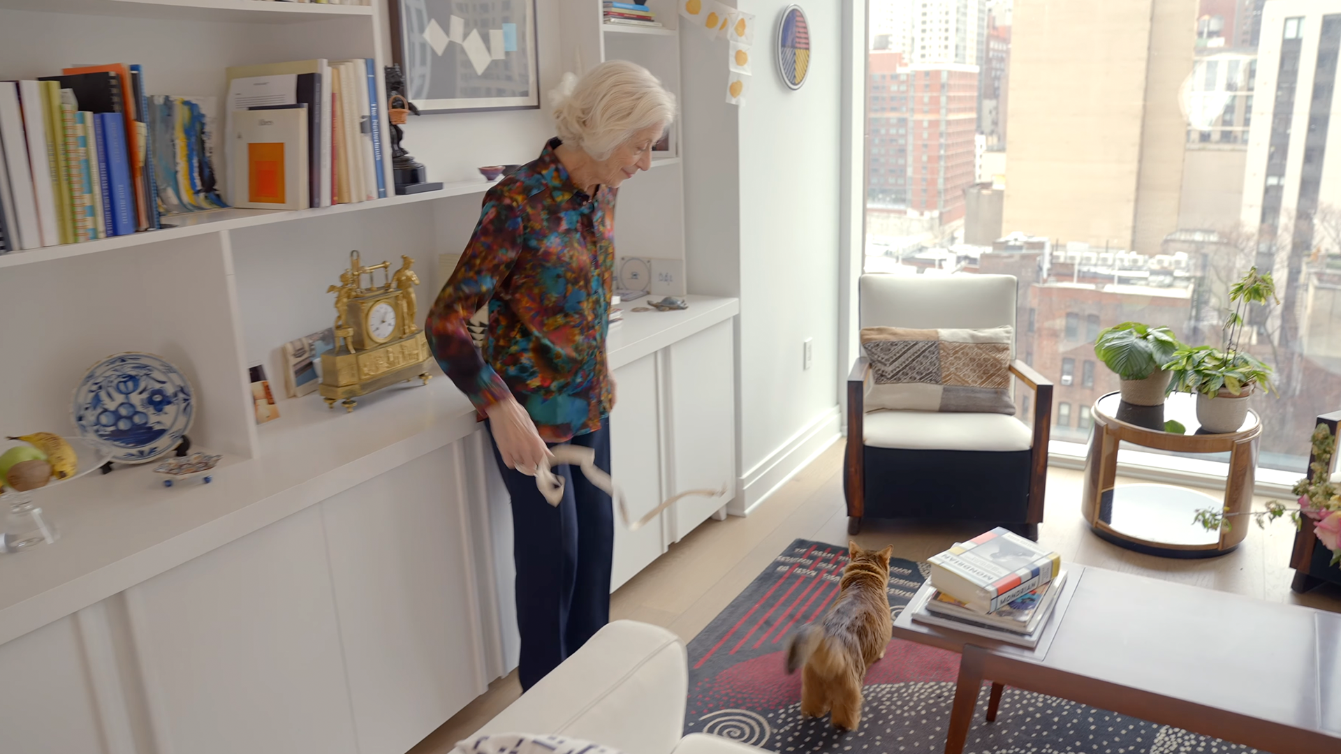 An elderly woman plays with a cat using a string in a modern, bright living room with bookshelves, plants, and large city-view windows.
