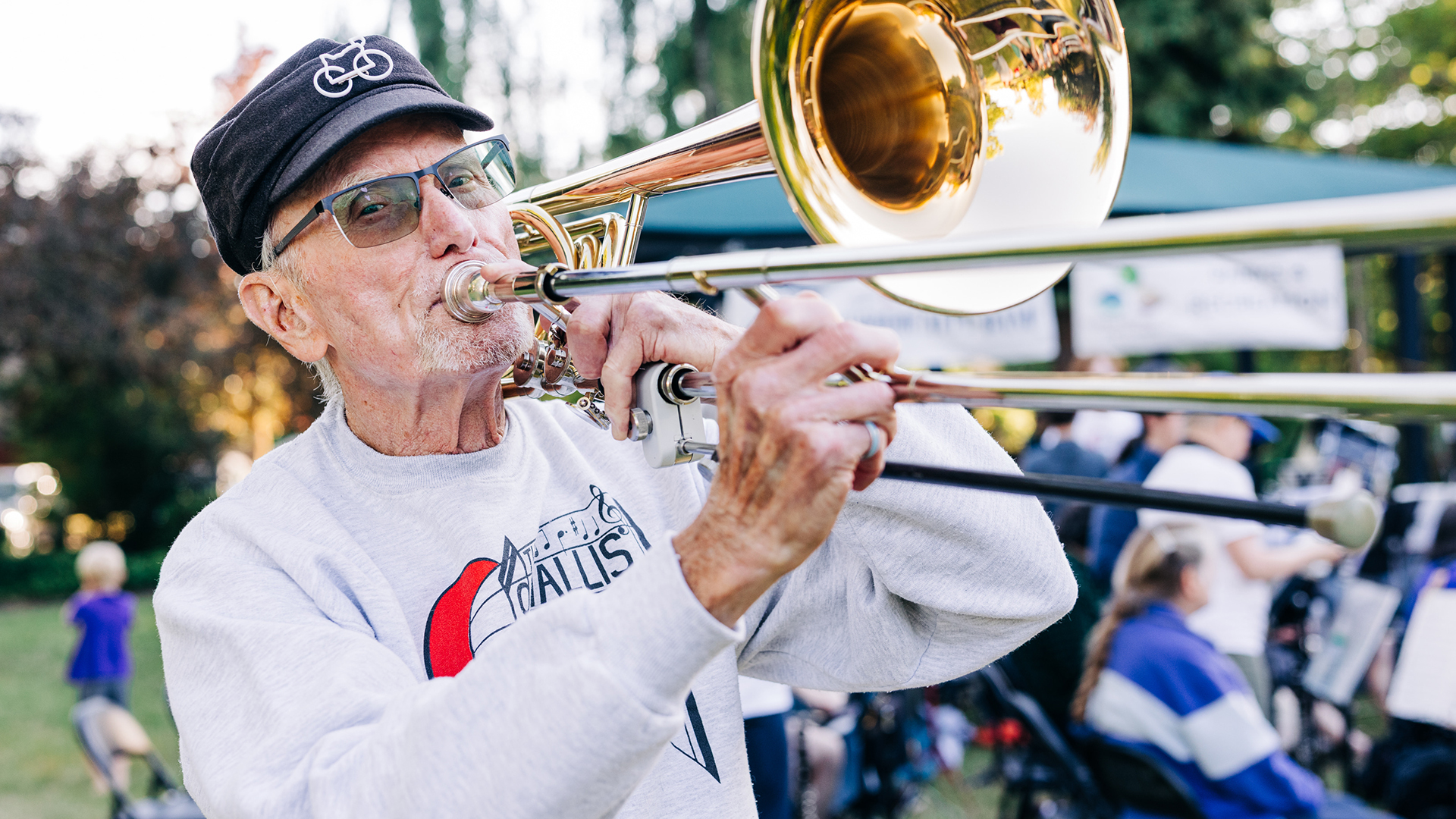 An older man wearing a cap and glasses plays a trombone outdoors, surrounded by people in a garassy area.