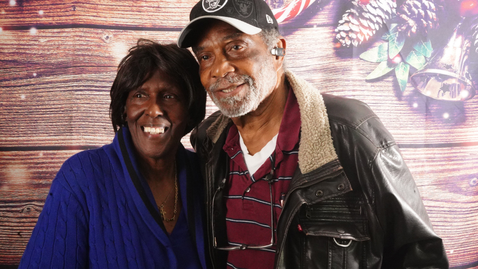 Older couple smiling warmly, standing close together in front of a festive wooden backdrop with pine cones and bells.