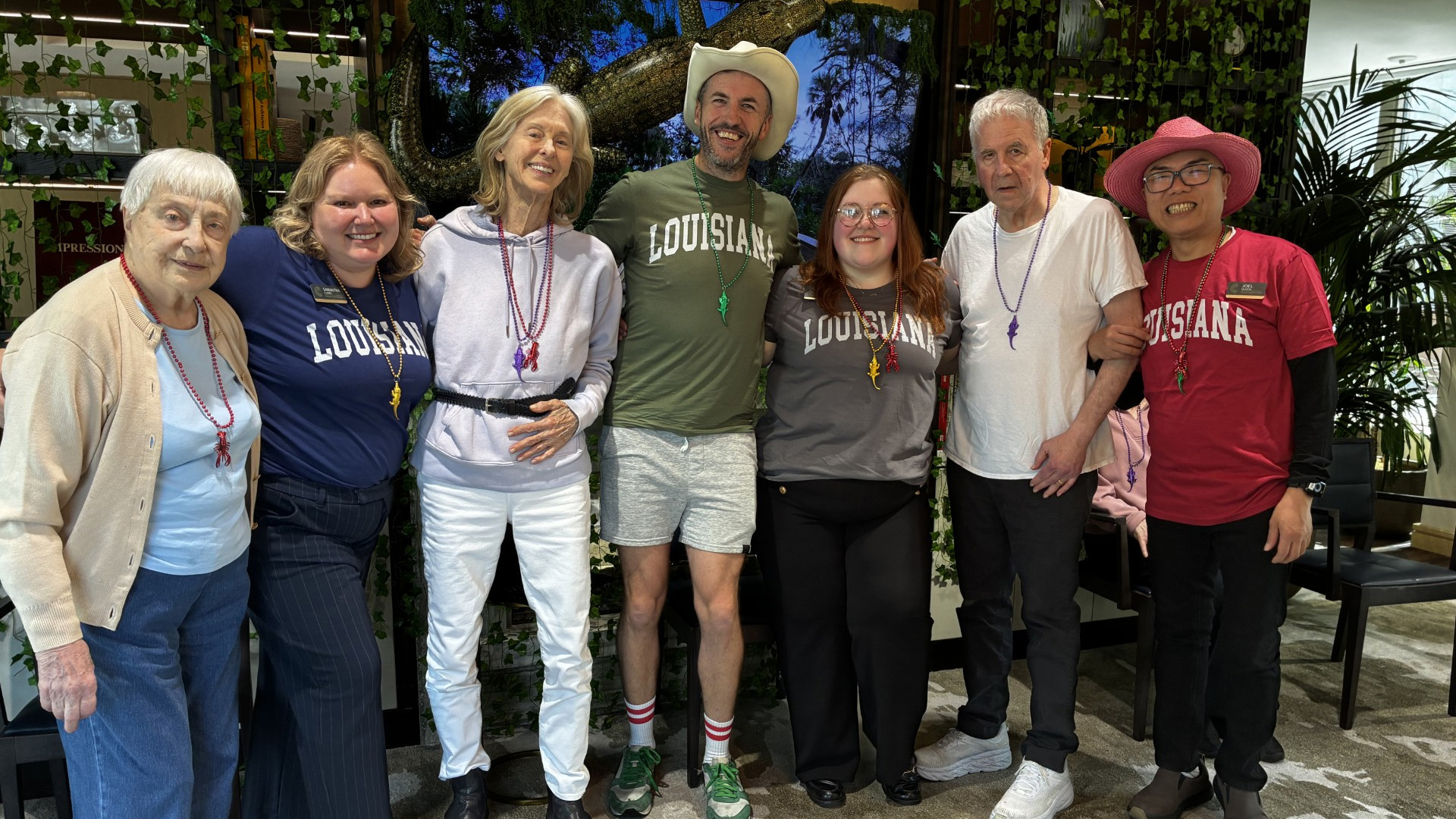 A group of seven people, wearing casual clothing and Mardi Gras beads, smiling and posing together in an indoor setting with greenery in the background.