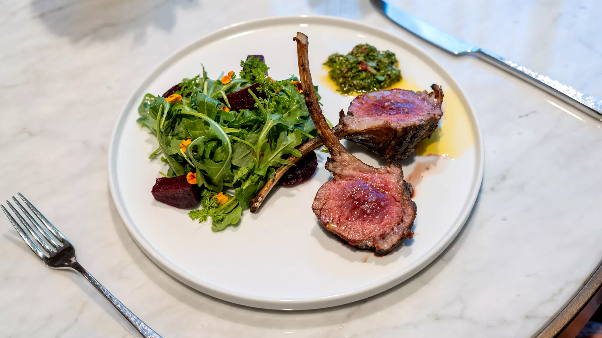 A plate of lamb chops with a side of arugula salad topped with edible flowers, beet slices, and a small serving of green chimichurri sauce.