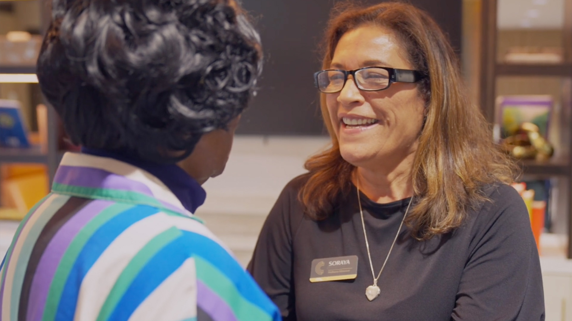 Two women smiling and talking; one wearing glasses and a nametag, the other in a colorful striped outfit.