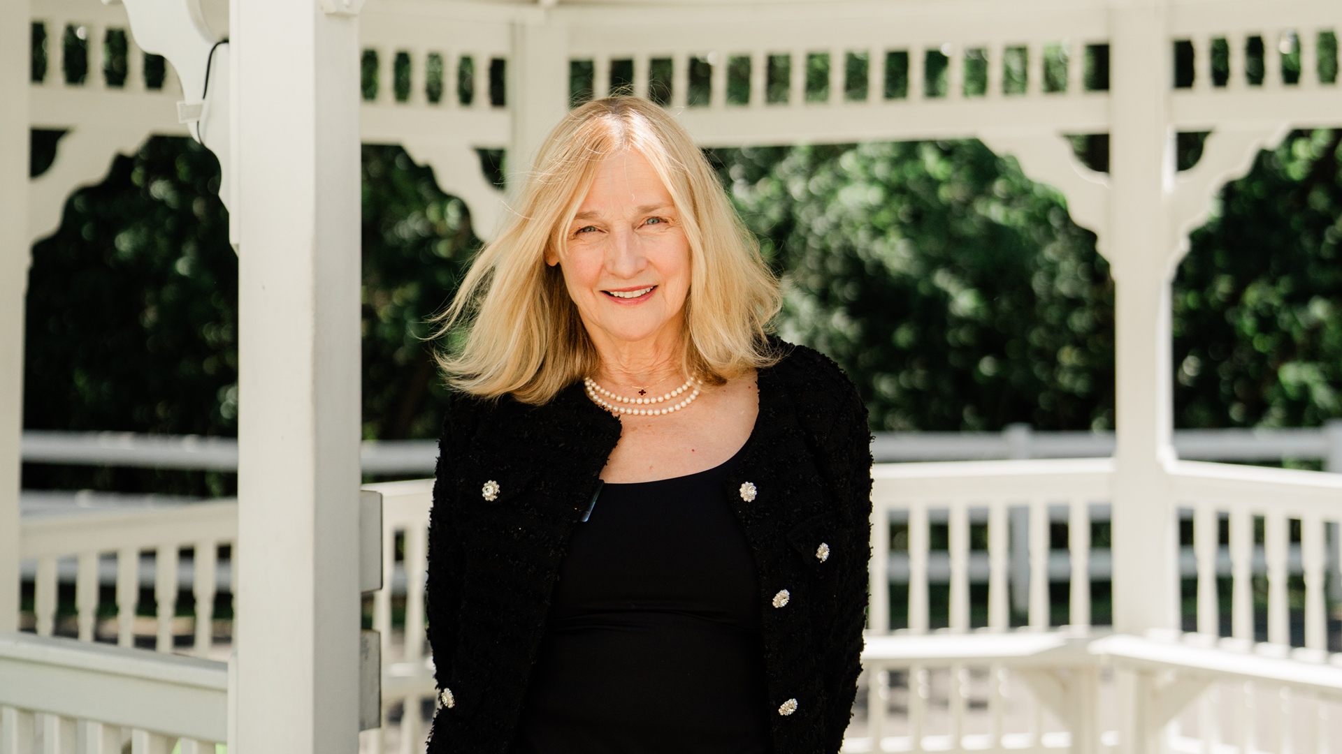 Woman with blonde hair and a black jacket standing in a white gazebo, smiling at the camera.