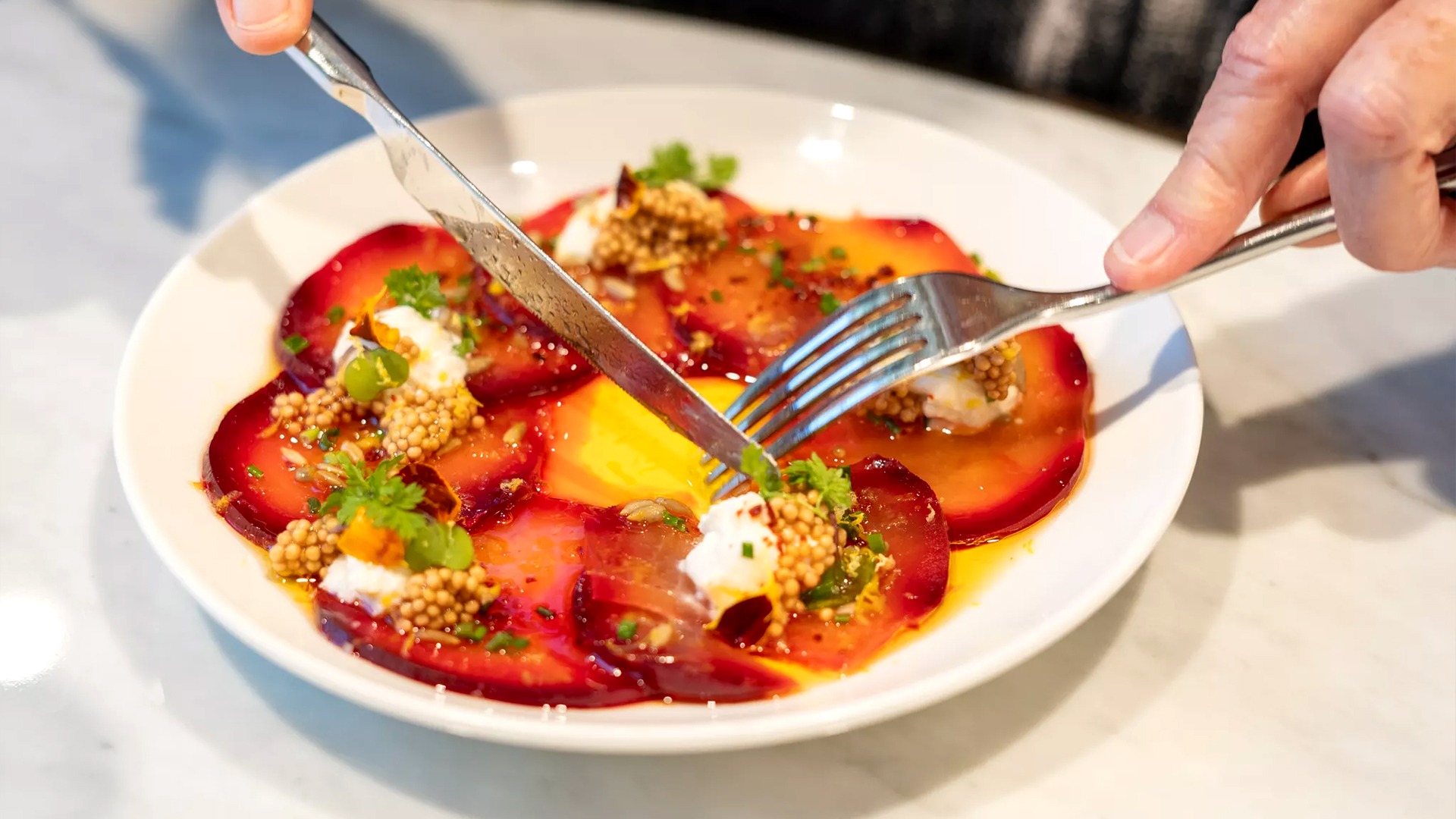 A hand holding a fork and knife cutting into a plate of vibrant beet slices garnished with mustard seeds, cream, and herbs.