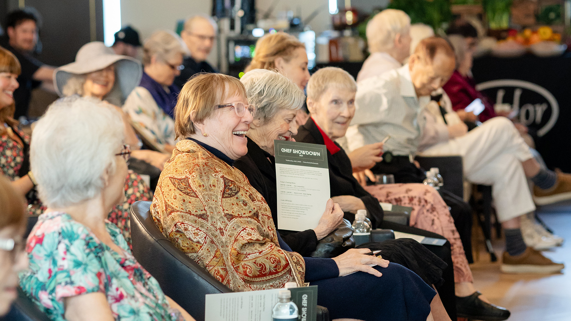 A group of older adults seated and laughing at an event, holding programs. The atmosphere is lively and joyful.