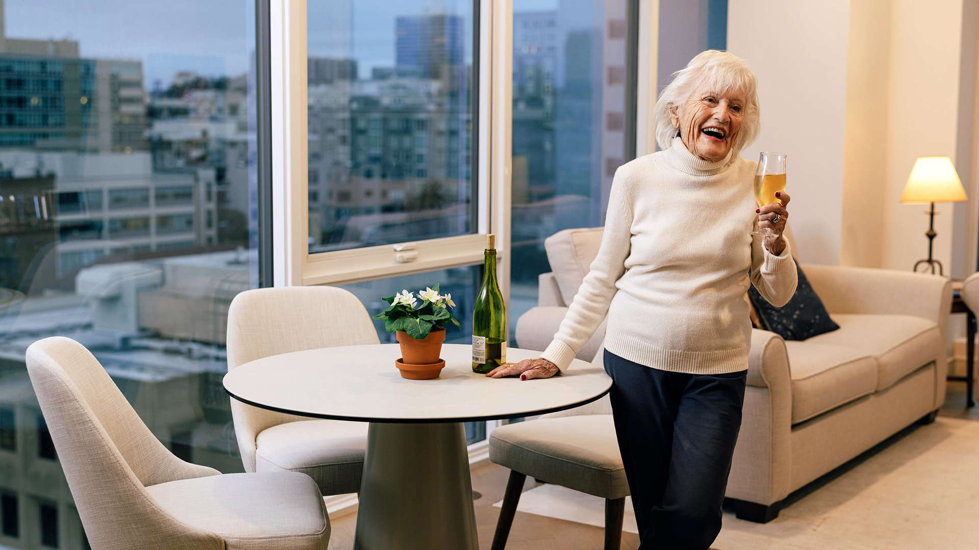 An older adult woman in a cozy apartment smiles while holding a glass of wine near a table with a potted plant and a bottle. City buildings visible outside.