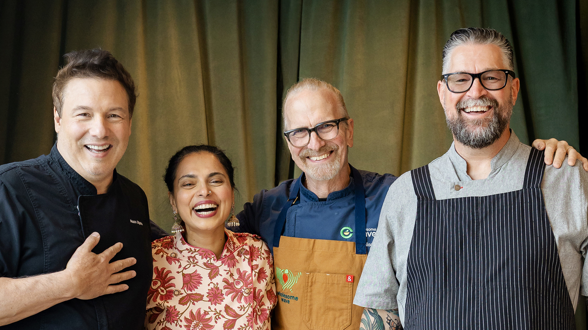 Four chefs smiling and standing together in front of a curtain, wearing various chef uniforms.