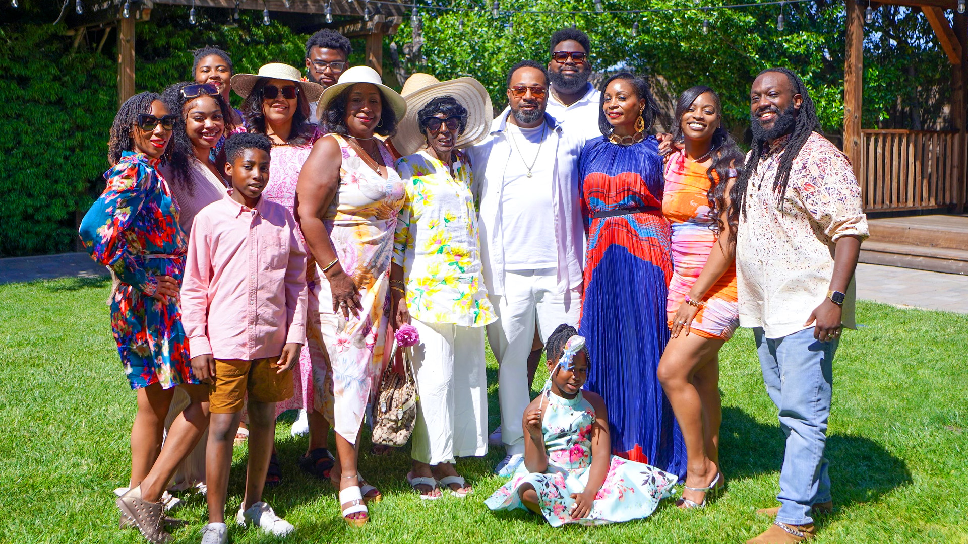 A diverse group of people posing together on a sunny day, wearing colorful summer outfits, with a girl sitting in front on grass.
