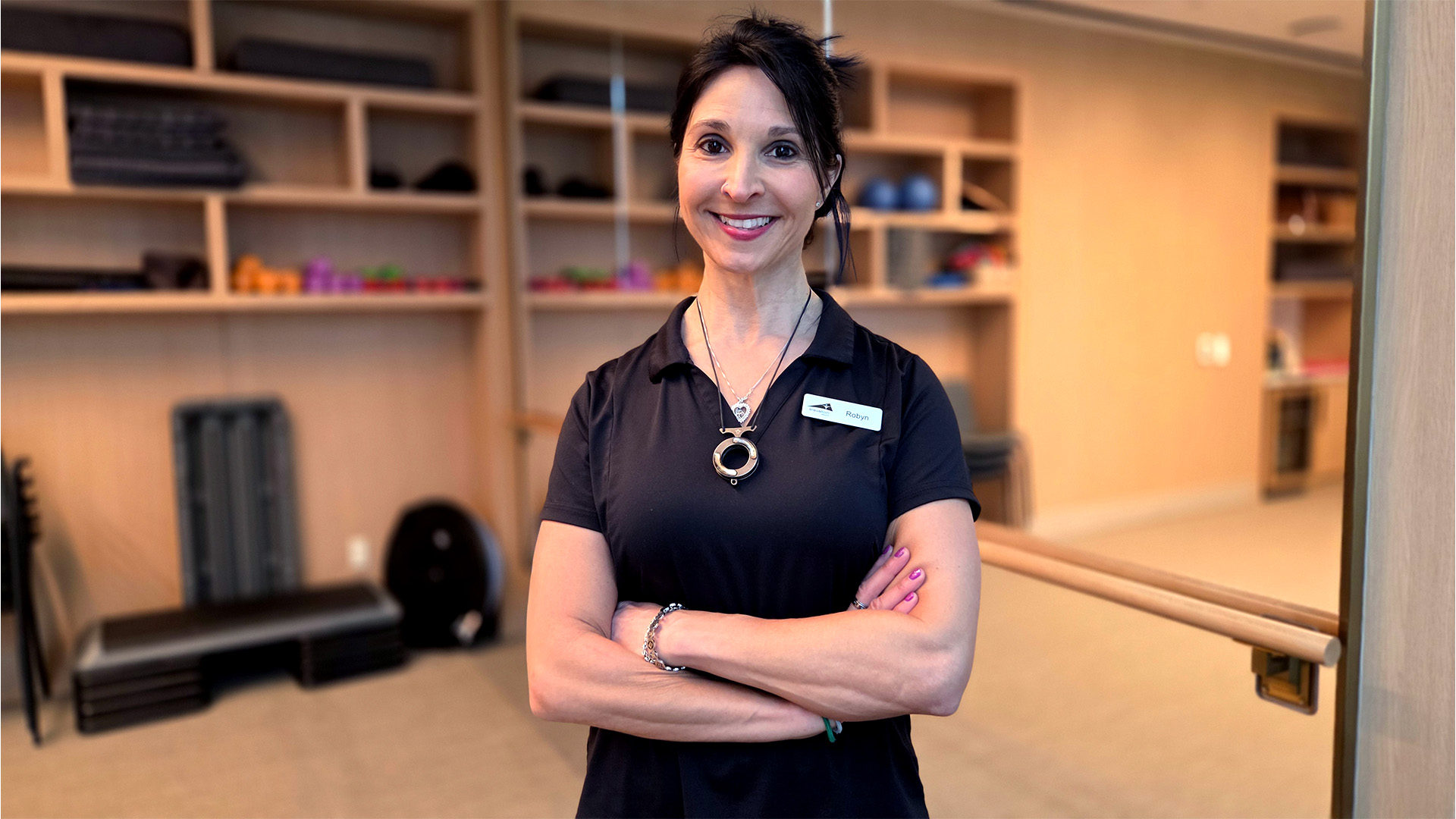 Woman smiling and standing with arms crossed in a fitness studio, wearing a black shirt and name tag. Shelves with exercise equipment in the background.