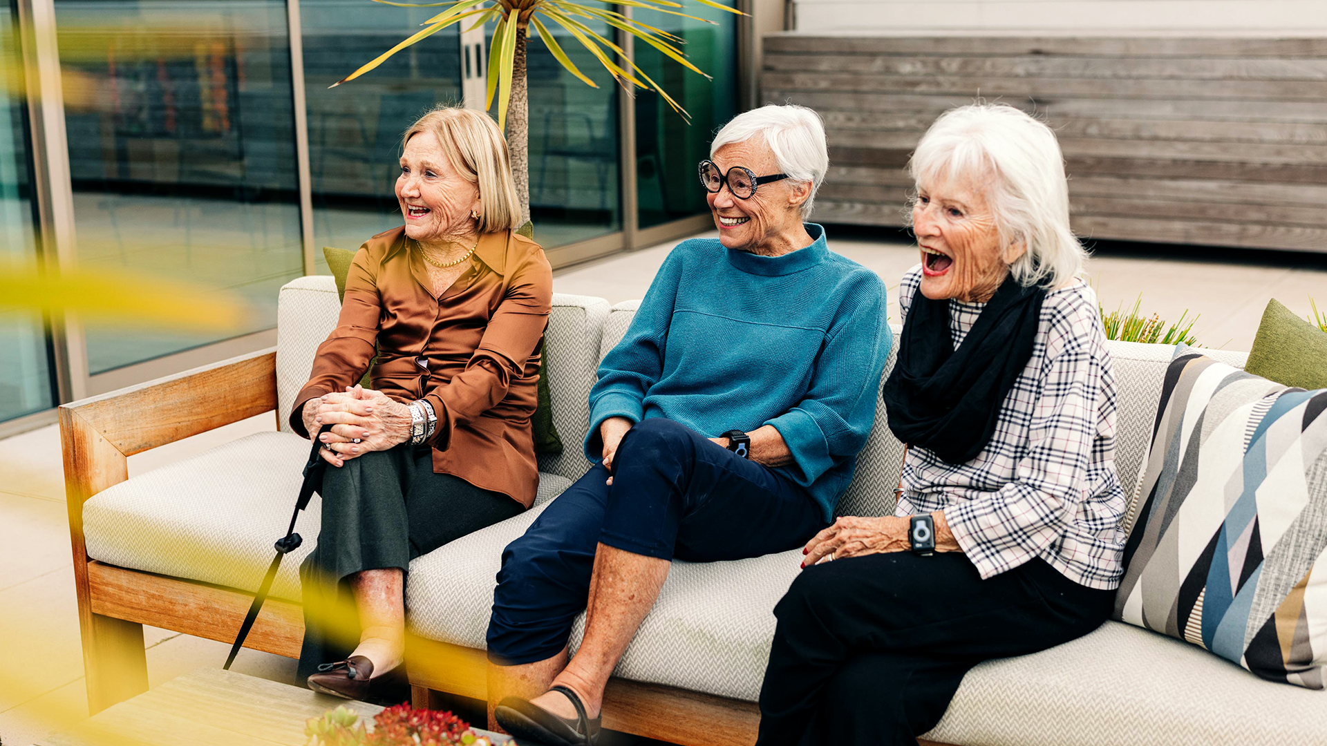 Three older adult women sitting on a patio couch, laughing and enjoying each other's company, with potted plants in the background.