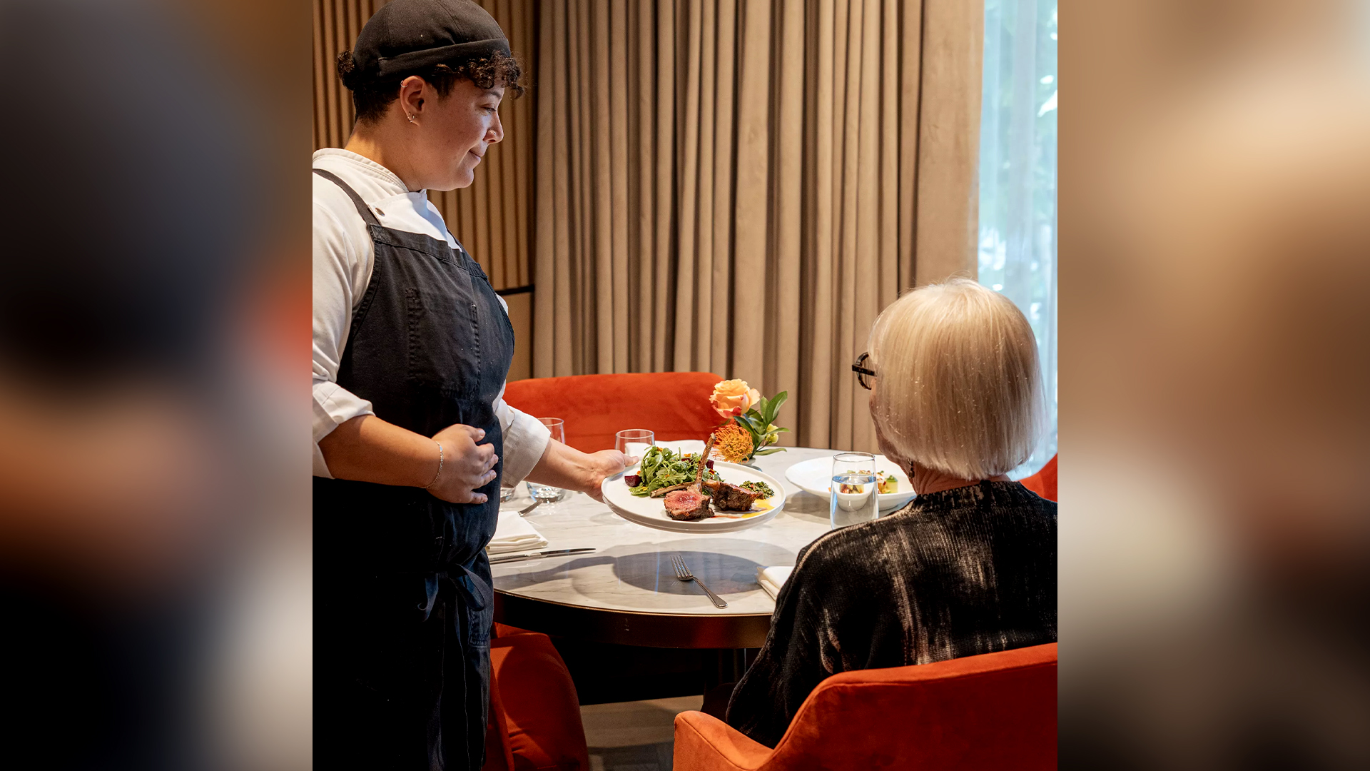 A server is presenting a plate of food to a seated woman in a stylish restaurant. The table is set with glasses and flowers.