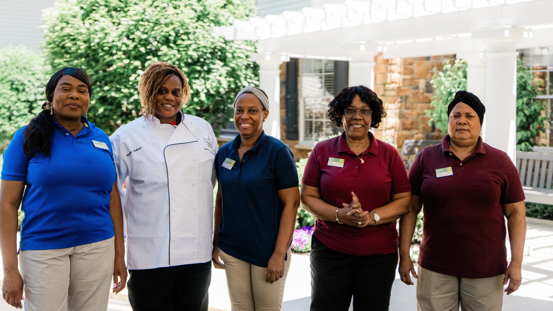 Five women, four in matching uniforms and one in a chef coat, smiling outdoors in front of a leafy background and a building.
