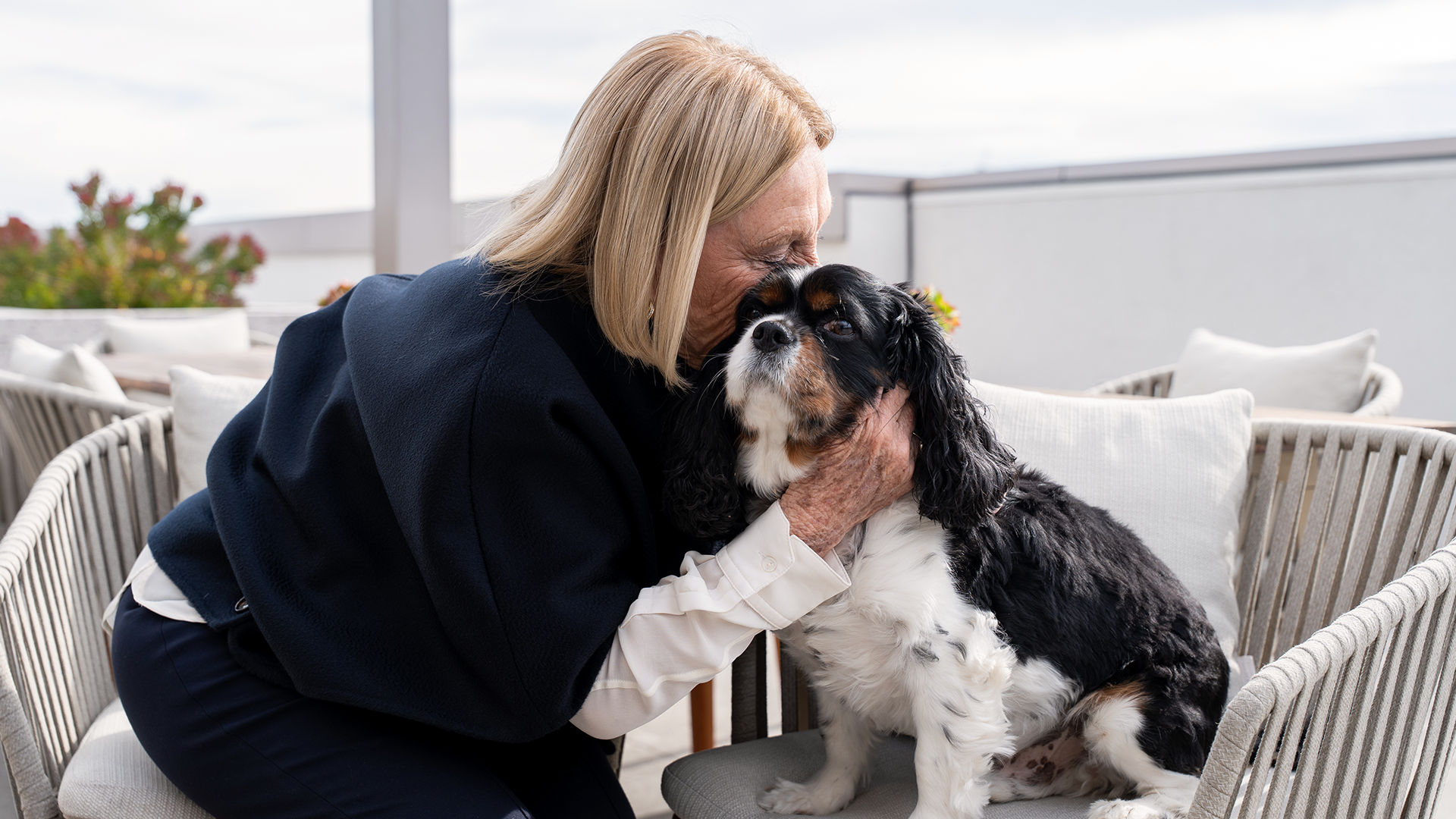 A woman with blonde hair lovingly cuddles a Cavalier King Charles Spaniel while sitting outdoors on a patio chair.
