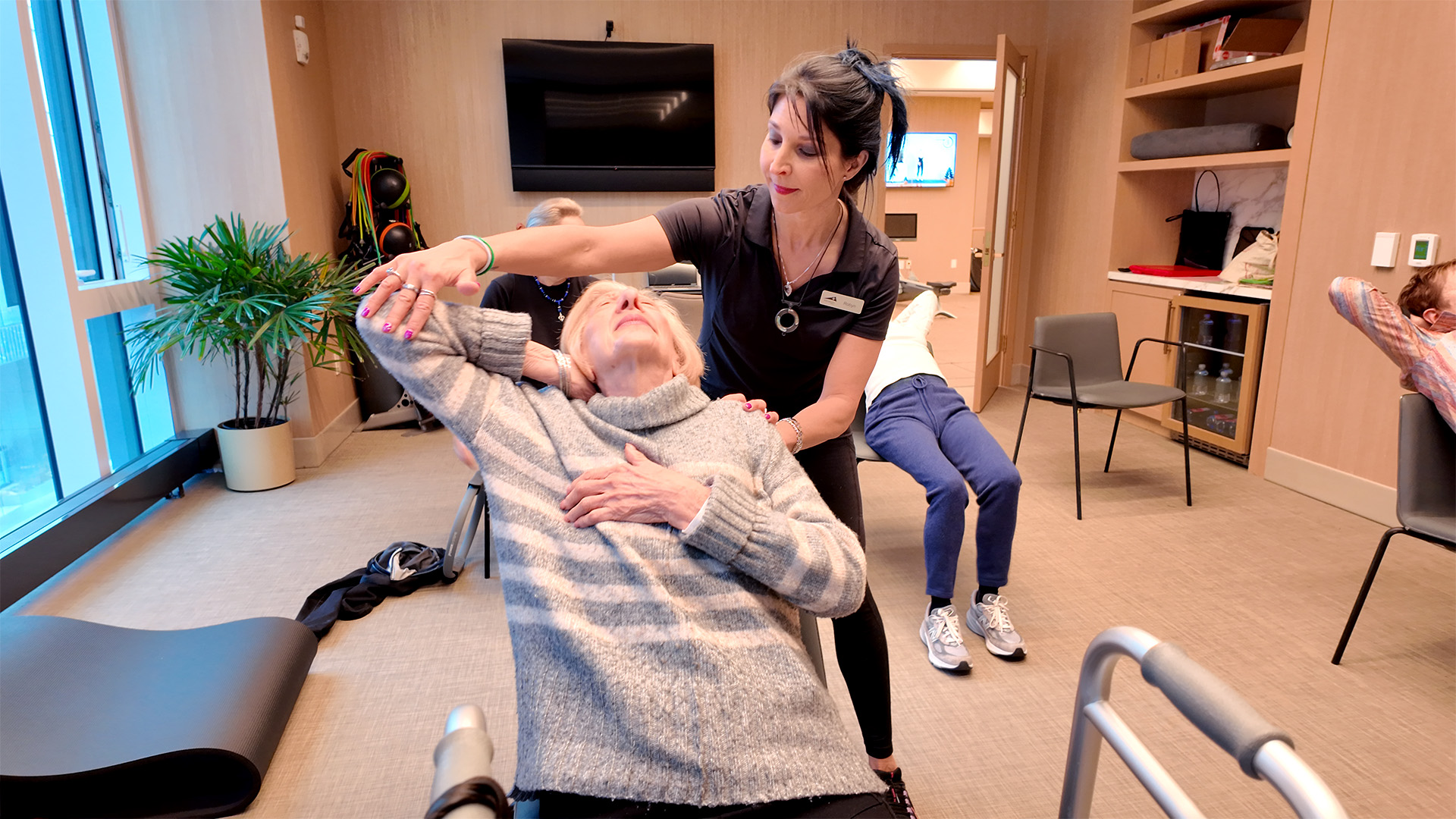 A woman assists an older woman with a stretching exercise in a bright, modern room equipped for physical therapy.