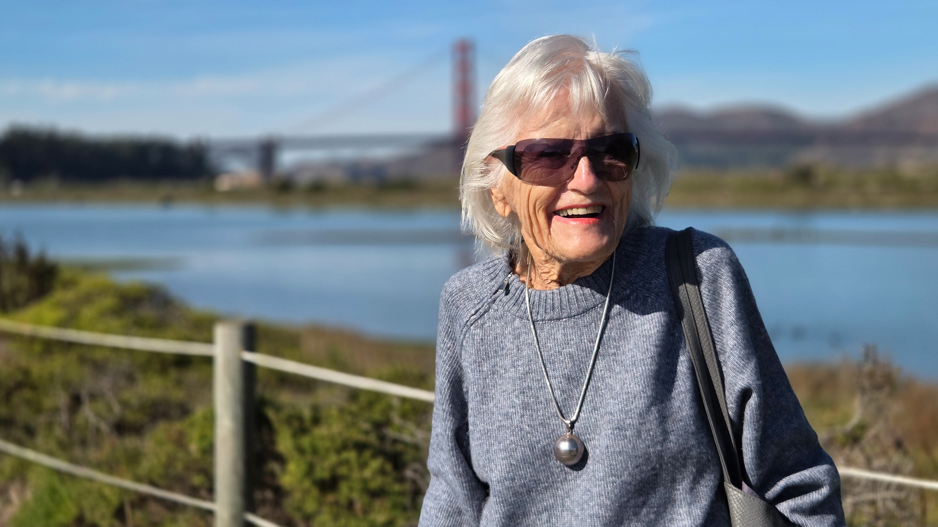 An older adult woman with white hair and sunglasses smiles outdoors, wearing a gray sweater and necklace. A bridge and water are visible in the background.