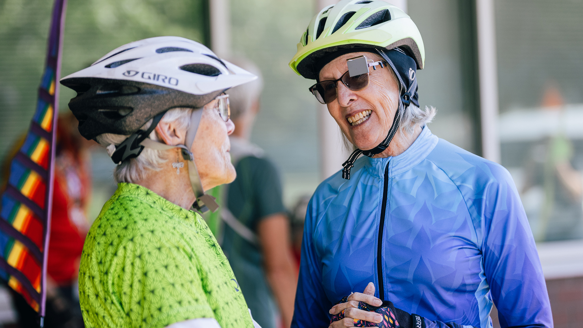 Two older women wearing helmets and cycling attire have a cheerful conversation outdoors, one in green, the other in blue, near a rainbow flag.