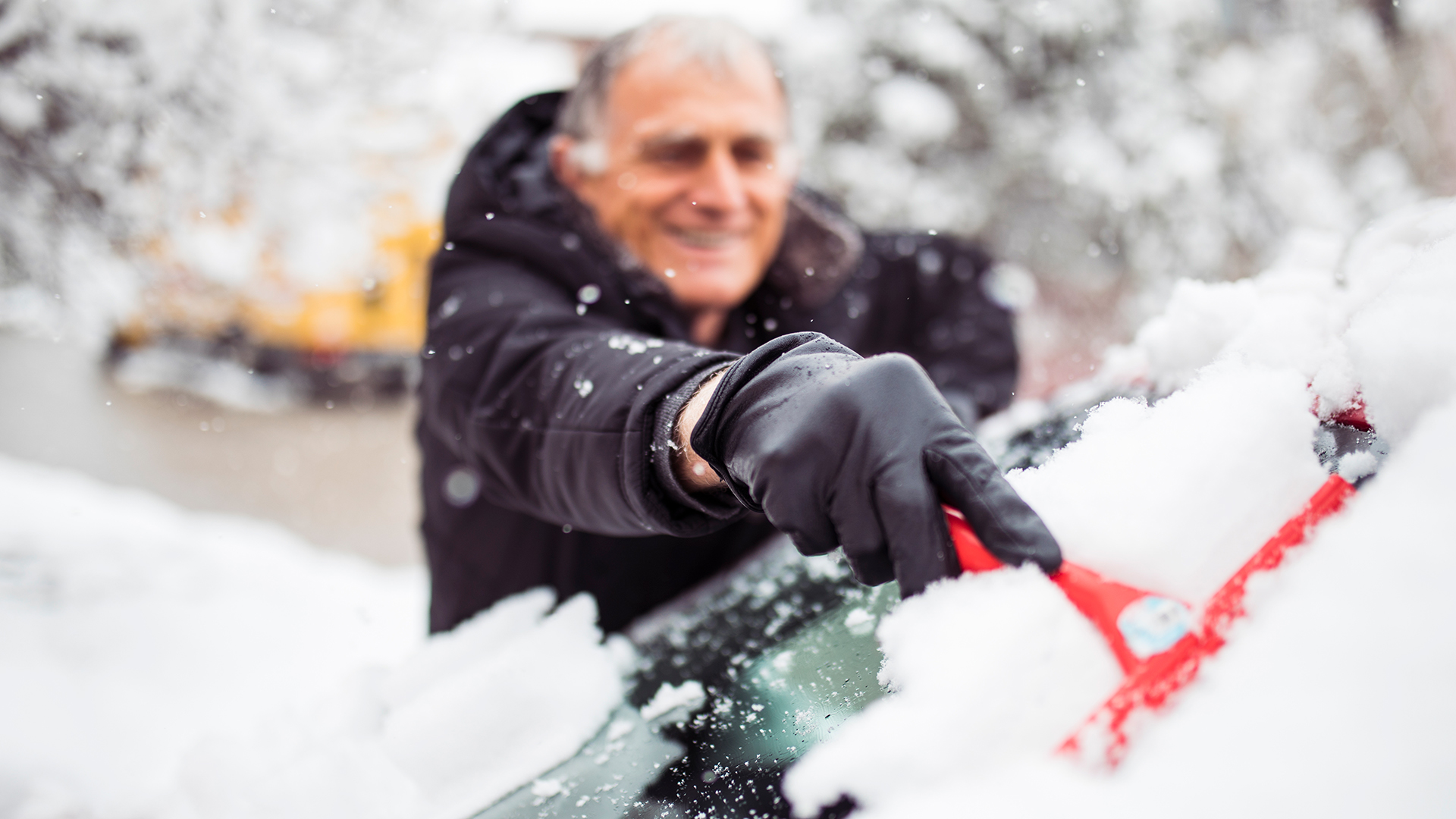 Man in a black coat and gloves scraping snow off a car windshield with a red brush as snow falls