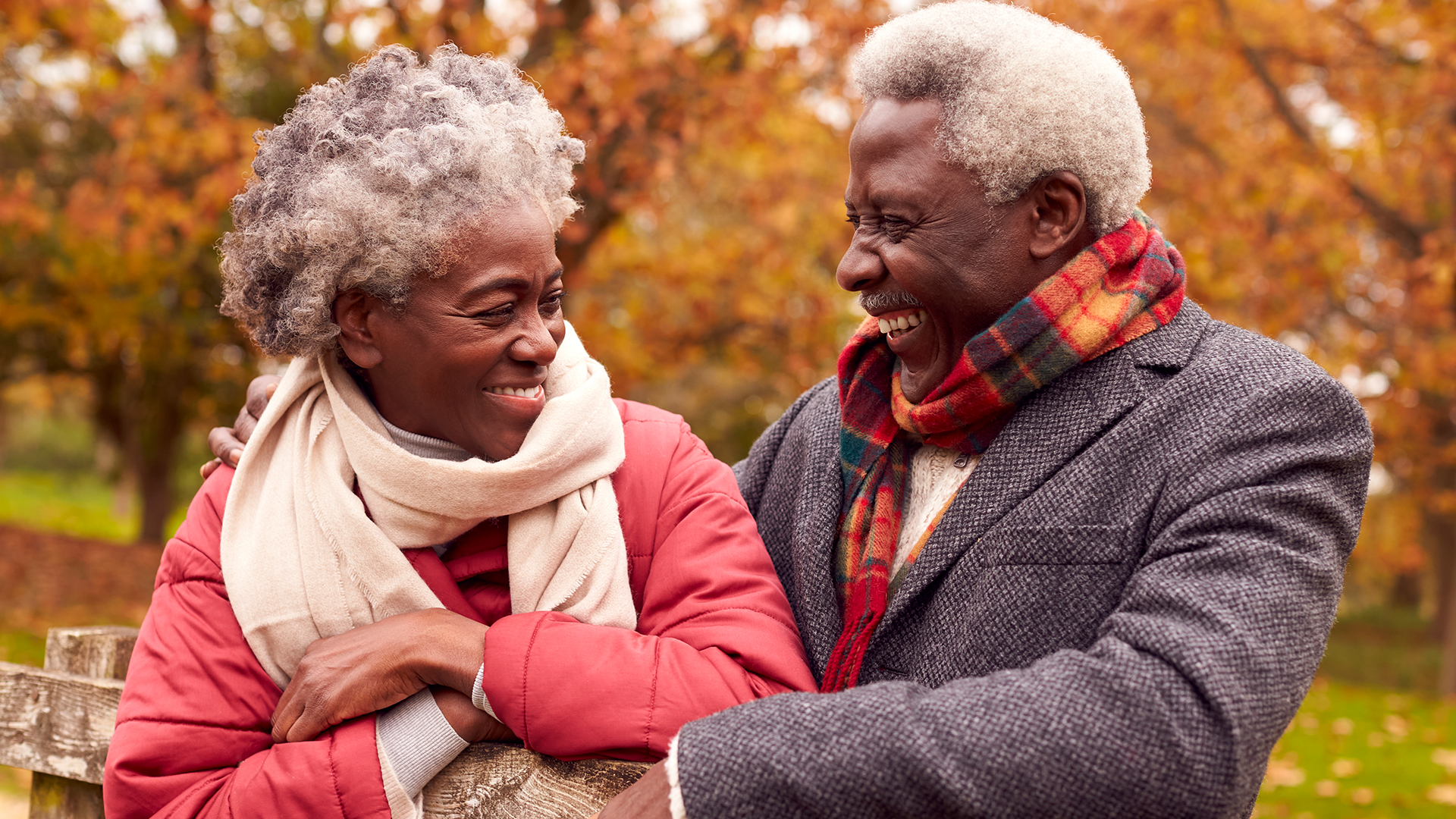 Smiling older couple in coats and scarves embrace on a park bench amid autumn foliage.