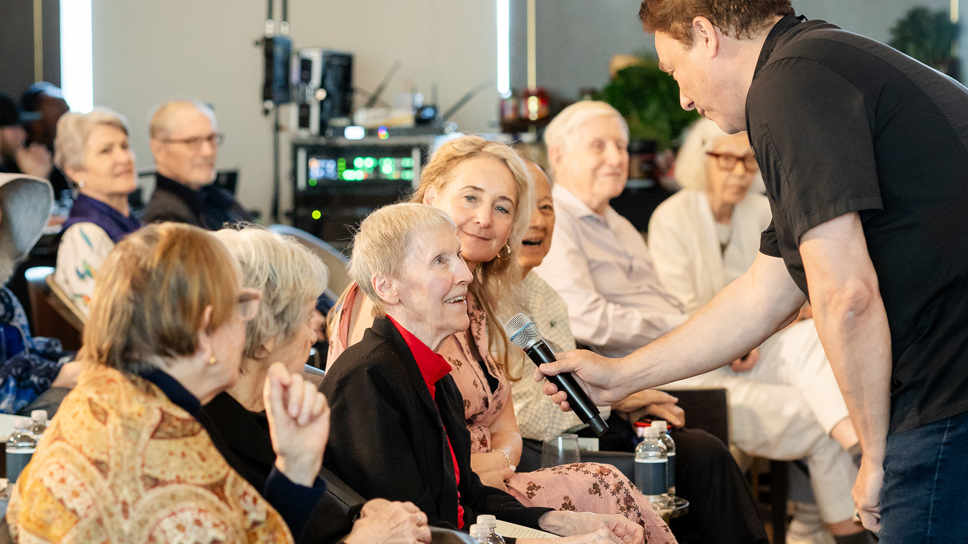 A man holds a microphone for an older woman in a seated audience, as others watch and smile in a well-lit room.