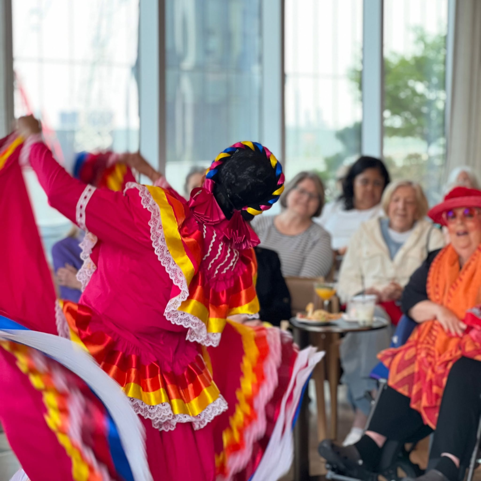 A dancer in vibrant traditional attire performs energetically in front of an appreciative audience seated indoors.