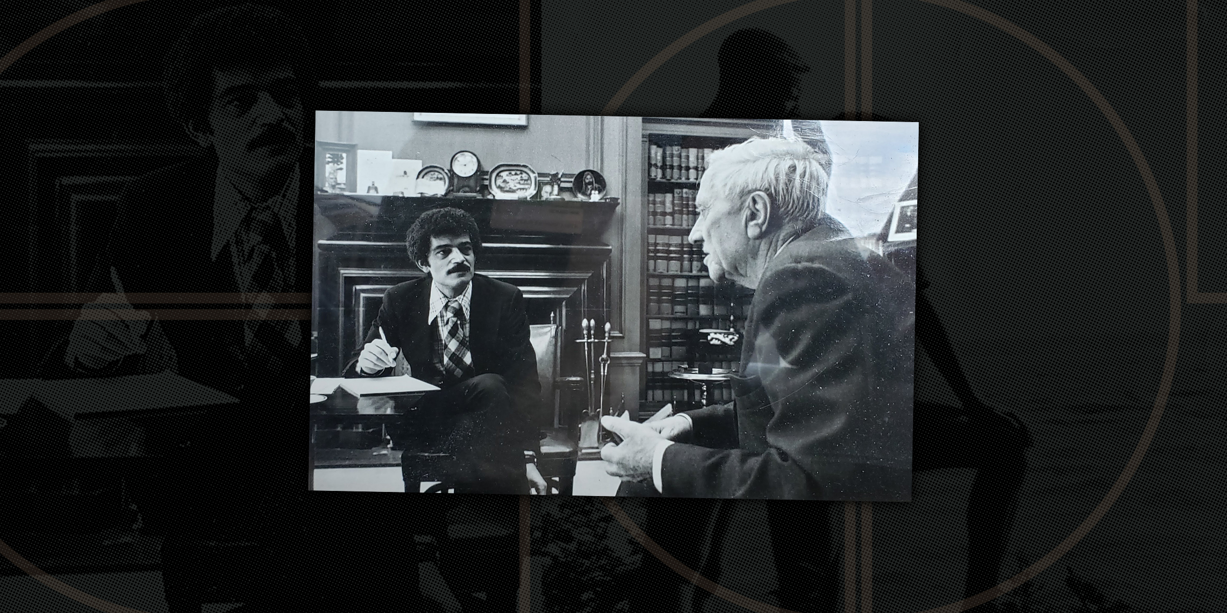 Black-and-white photograph of two men in a formal office setting. One man, seated at a desk and holding a pen, listens attentively while the other older man, seated across from him, speaks. The room has wood-paneled walls, shelves filled with books, and a mantel displaying framed items.