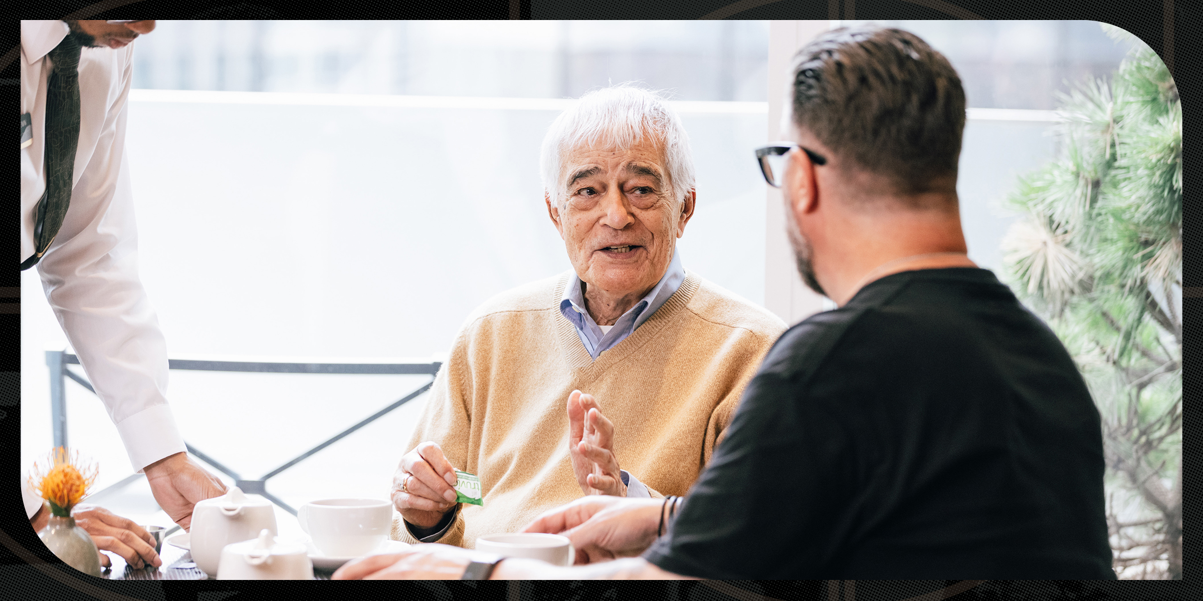 An older man sits at a table in a bright café, speaking with another man seated across from him. A server reaches in to place a teapot on the table. The older man holds a packet of sweetener and gestures mid-conversation. Cups, teapots, and a small vase with a flower are on the table, with large windows and greenery in the background.