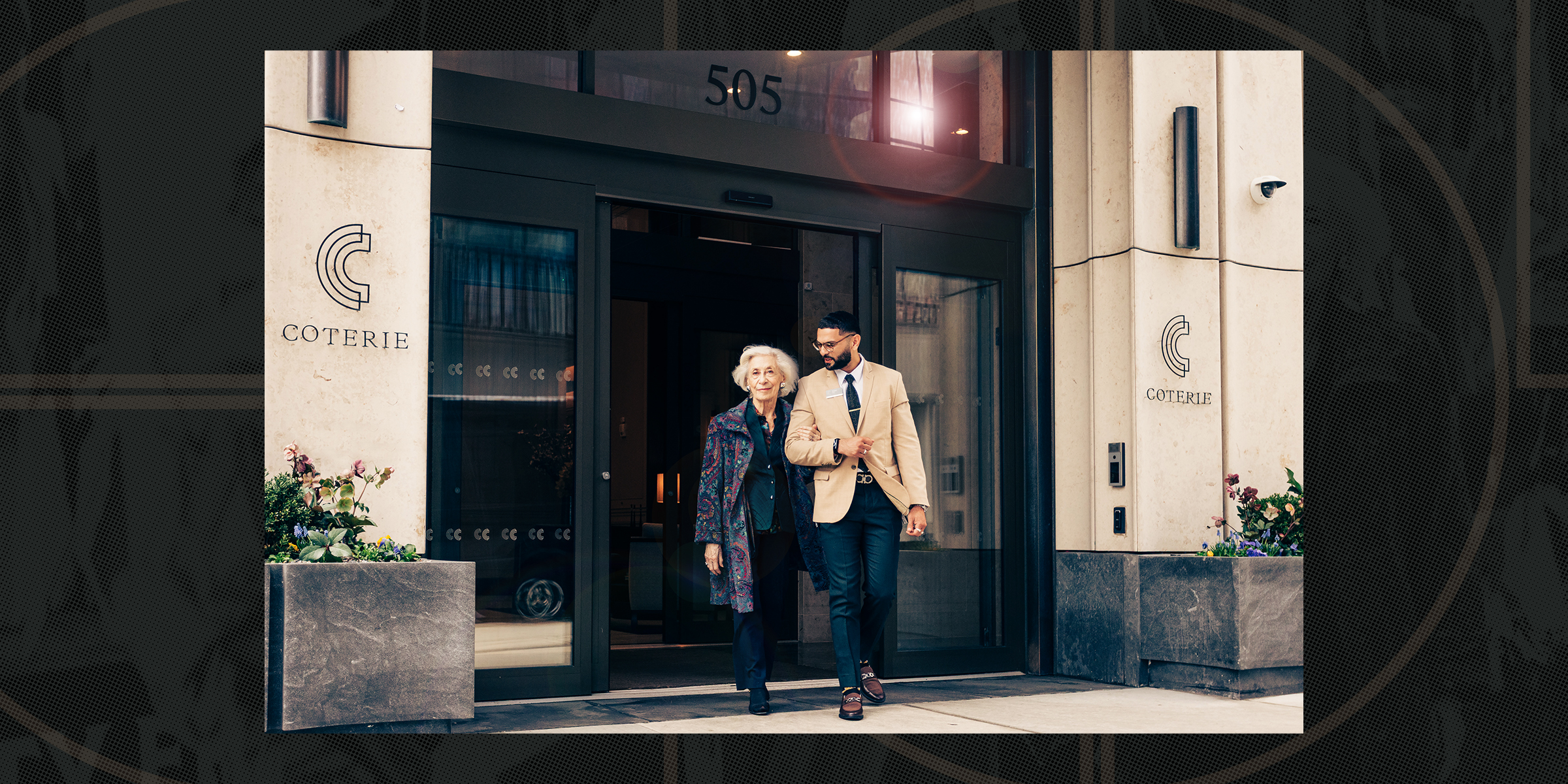 An older woman and a younger man walk arm-in-arm, smiling, outside a building labeled 