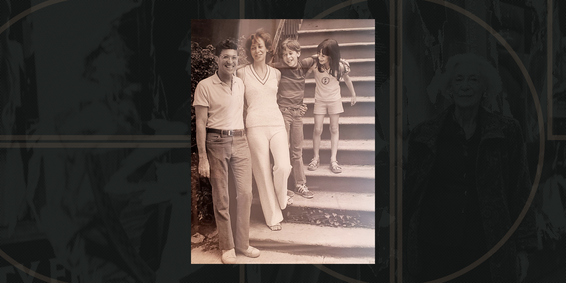 A family of four, two adults and two children, stand smiling on outdoor steps. The adults are casually dressed, and the children lean playfully.