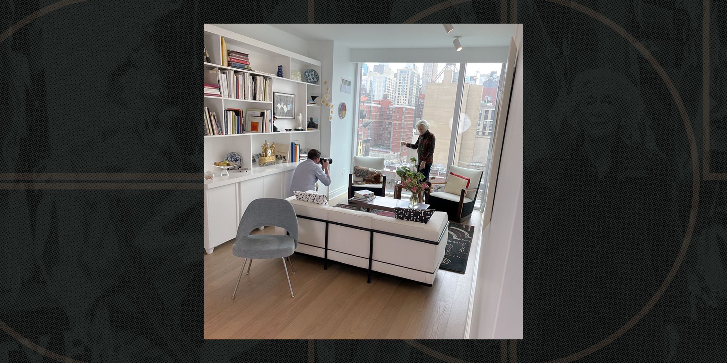 A photographer takes pictures of a woman in a stylish living room with shelves, books, and a cityscape view through large windows.