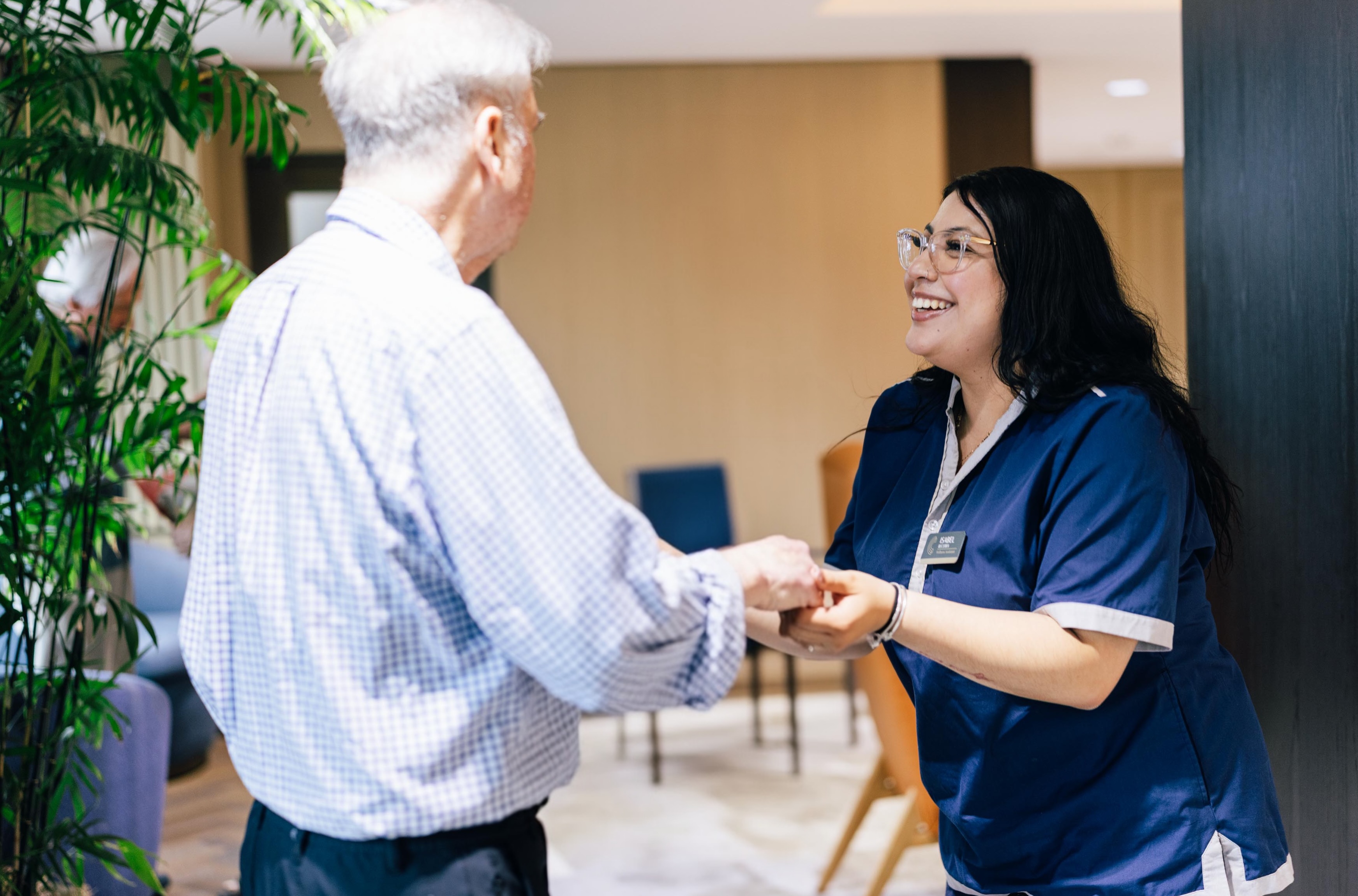 A smiling woman in a blue uniform warmly holds hands with an older man in a checked shirt in a well-lit indoor space.