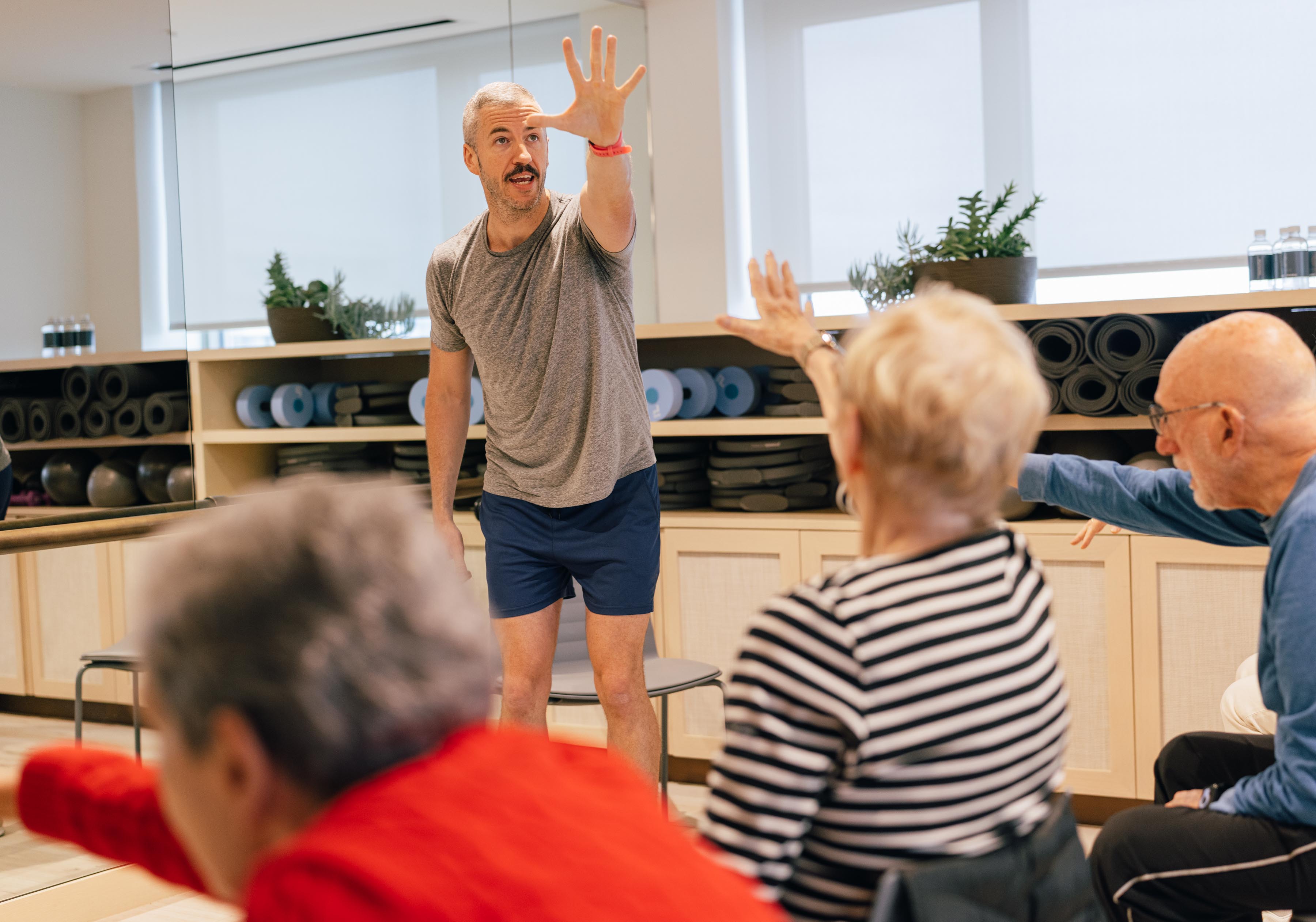 A fitness instructor demonstrates an exercise move to a group of older adults in a class, with yoga mats and exercise balls in the background.