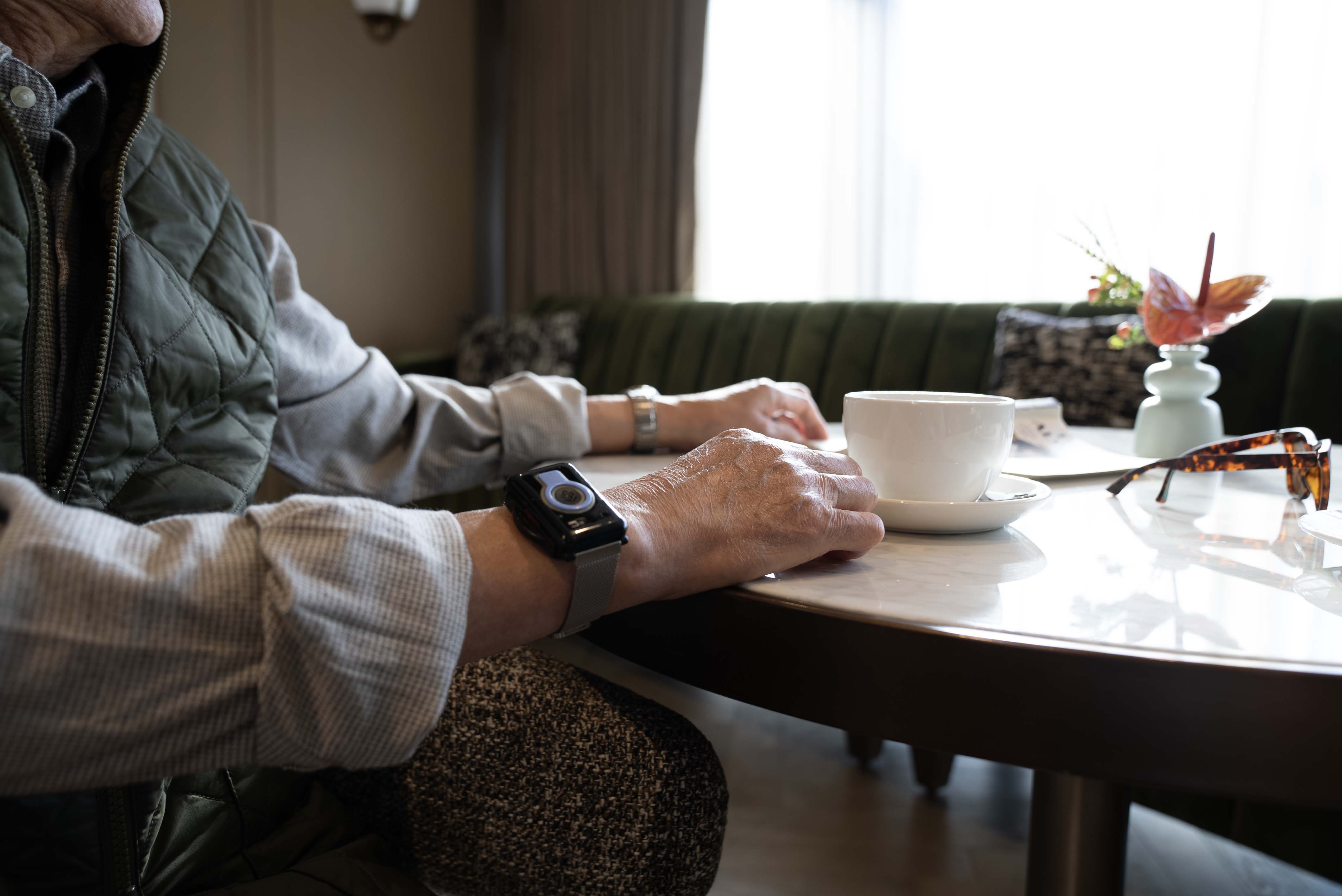 An elderly person wearing a smart wrist device sits at a table with a cup of coffee, glasses, and a vase, in a cozy, softly lit room.
