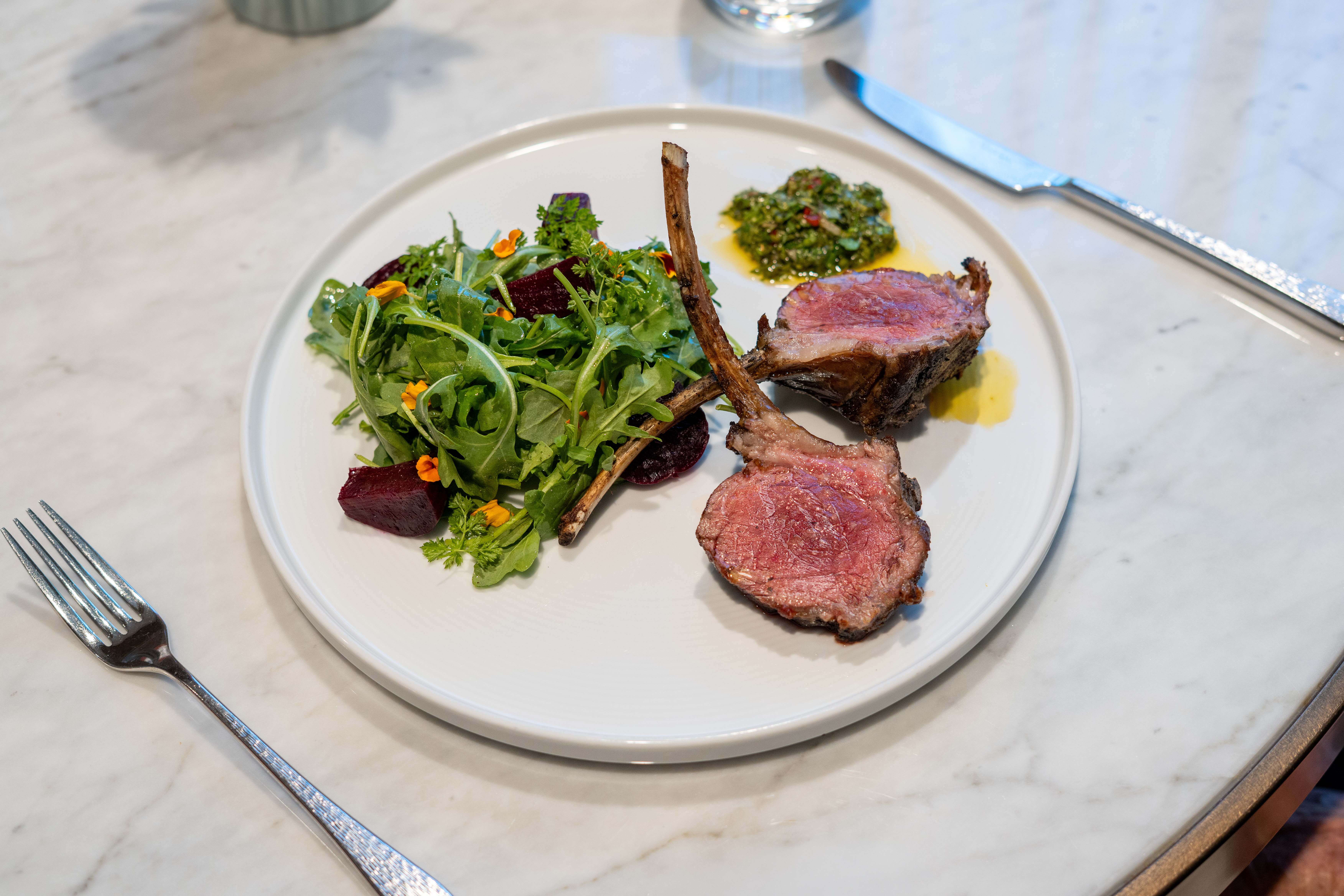 A plate of lamb chops with a side of arugula salad topped with edible flowers, beet slices, and a small serving of green chimichurri sauce.