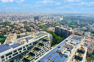 Aerial view of a cityscape featuring buildings with solar panels on rooftops, surrounded by greenery and dense urban development under a blue sky.